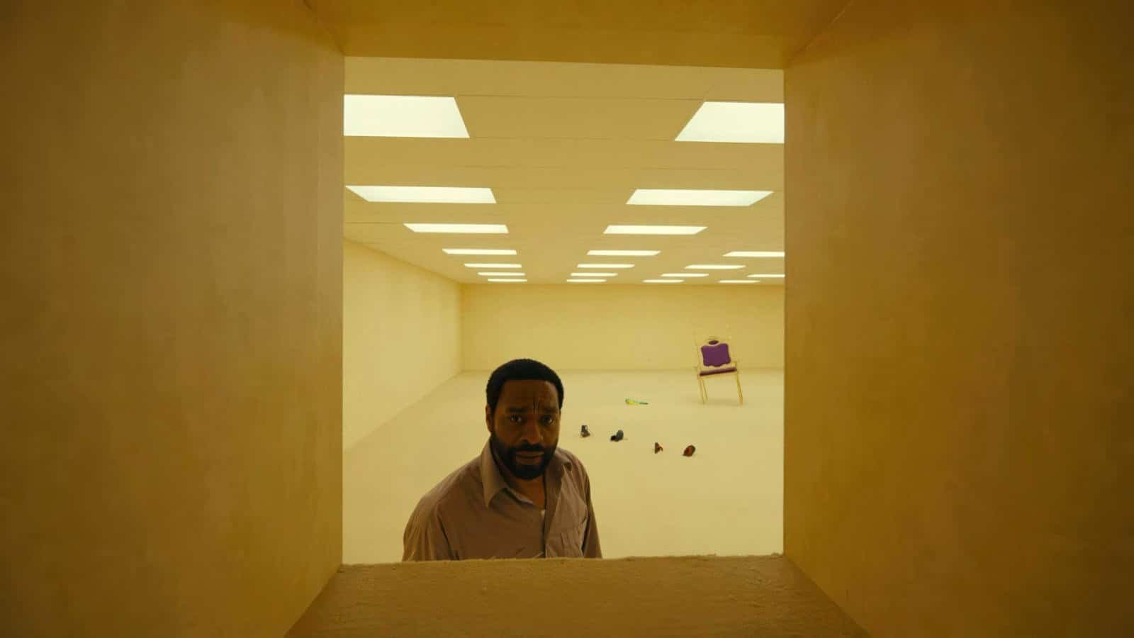 A Black man (Chiwetel Ejiofor) in a button-down shirt stands in a room with yellow wallpaper and a scattering of random objects on the floor behind him and peers through a chest-high, square opening in the wall in Backrooms