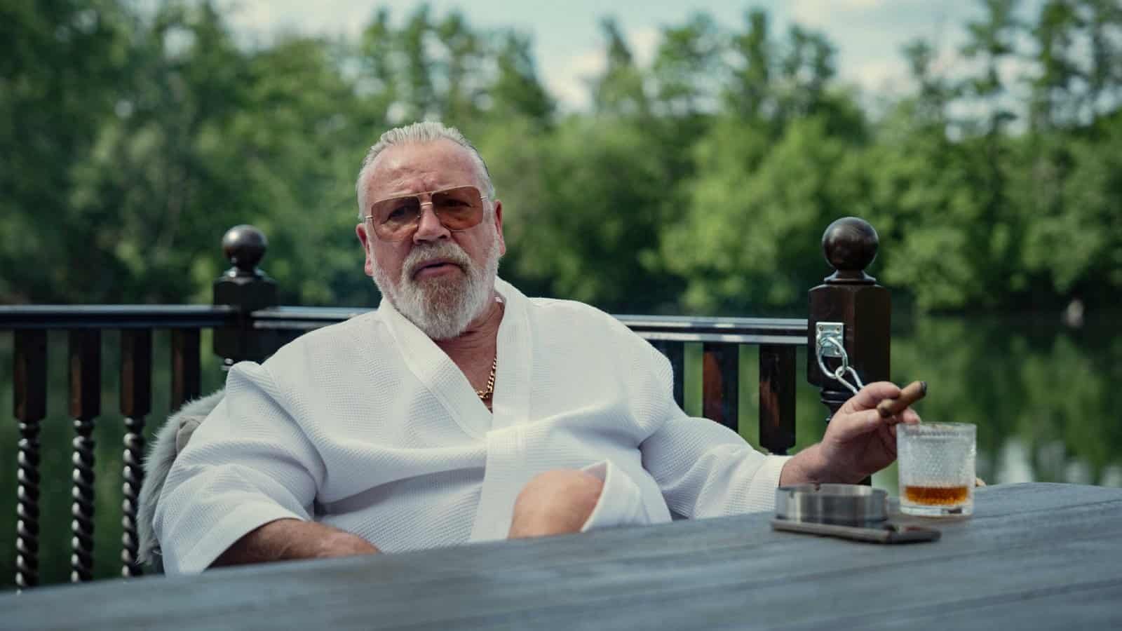 Bobby Glass (Ray Winstone) sits with a drink outside with a lake behind him in The Gentlemen season 2